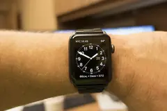 Черен Apple Watch с метална верижка на китка, на екрана аналогов циферблат с бели стрелки
